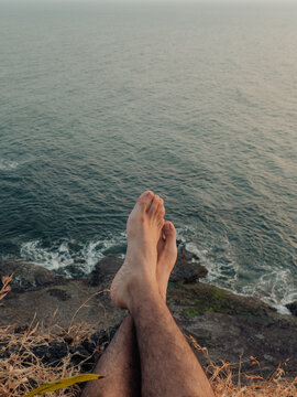 View of bare feet dangling over a rugged cliff edge, where golden grass meets the turquoise sea in a serene coastal moment, Chiplun, Maharashtra, India.