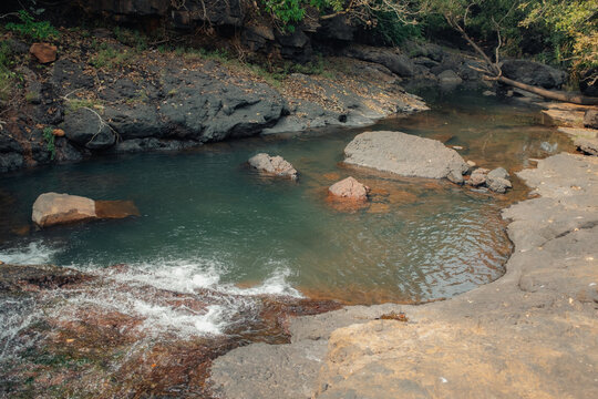View of a serene stream flowing between rocky banks, with sunlight glinting on the water's surface, creating a tranquil scene, Chiplun, Maharashtra, India.