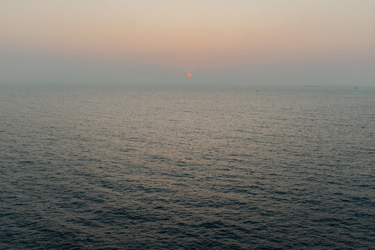 View of the sun's crimson orb sinking into the tranquil, dark waters of the Arabian Sea under a hazy sky, casting a serene glow, Chiplun, Maharashtra, India.