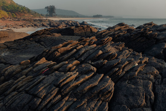 View of rugged, dark rocks meet the shoreline as gentle waves roll onto a sandy beach, framed by distant hills under a soft sky, Chiplun, Maharashtra, India.