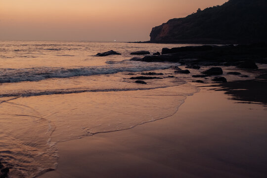 View of the tranquil beach meeting the ocean under a dusky sky, with rocks scattered along the shore, creating a serene coastal scene, Chiplun, Maharashtra, India.