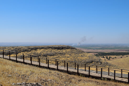 View of a wooden walkway leading across a golden, grassy hilltop under a vast blue sky, with distant smoke plumes adding depth, Urfa, Sanliurfa, Turkey.