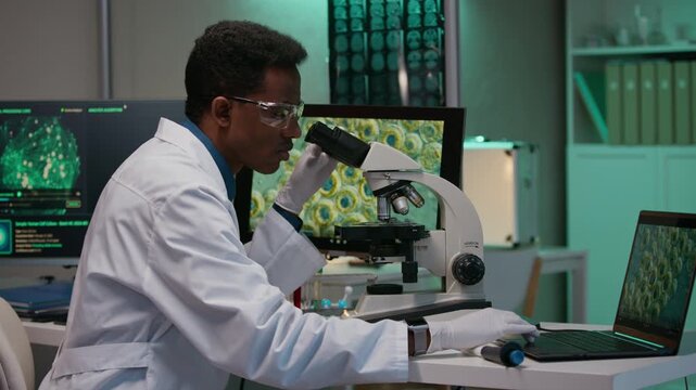 Side view of young male Black researcher in white coat examining cells under microscope and using smart software and applications on digital devices in clinic laboratory