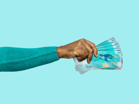 A hand holding a fanned stack of Maldivian 1,000 Rufiyaa banknotes against a solid blue background. The vibrant polymer bills feature a sea turtle design, symbolizing wealth and Maldivian culture.