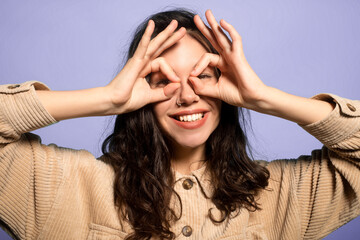 A young person stands against a light purple background. They smile while using their hands to create a playful gesture around their eyes. Their hair is dark and wavy. © Prostock-studio