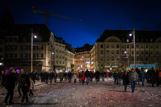 Basel, Switzerland - 10 March 2025: View of a crowded square shimmering under the night sky, littered with confetti, surrounded by the dark silhouettes of grand buildings.