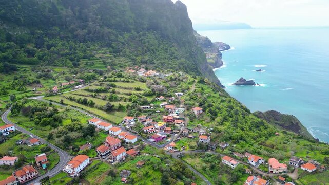 Aerial view of a vibrant Arco de Sao Jorge village nestled on the lush green cliffs of Madeira, with the Atlantic Ocean stretching to the horizon under a cloudy sky.