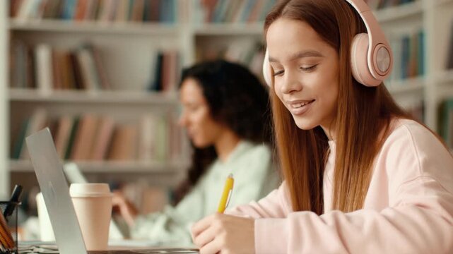 Girl with light brown hair wears headphones while taking notes in a library. She smiles as she studies at a laptop with her classmate nearby also working.