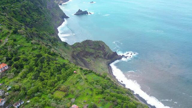 Dramatic aerial view of Madeira's lush green coastal cliffs meeting the vibrant blue Atlantic, with waves crashing on a dark pebble beach. Arco de Sao Jorge