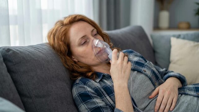 Unwell red haired woman with breathing problems using a nebulizer for inhalation treatment, lying on the couch at home for respiratory therapy against asthma or bronchitis