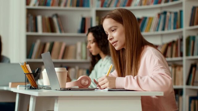 A young girl with long brown hair is focused on writing notes at a study table in a library. Two other students work on laptops in the background. The library has rows of bookshelves.