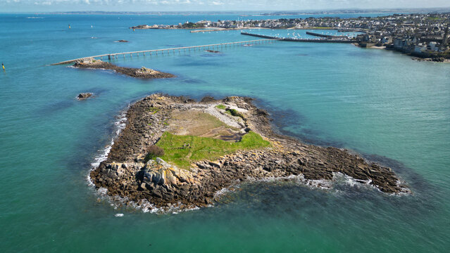 Aerial view of rocky islands surrounded by turquoise waters, connected by a long pier stretching towards the distant town, Roscoff, Brittany, France.