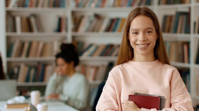 A young woman with light skin and long brown hair smiles while holding notebooks in a library setting. Behind her, another woman reads quietly at a table. Bookshelves surround them.