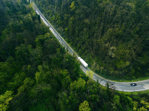 Aerial view of a truck and car navigating a winding road snaking through dense forest, creating a striking contrast between nature and transport, Novi Sad, Vojvodina, Serbia.