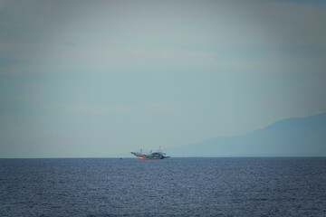 Obraz premium A lone fishing boat drifts across open water with hazy mountains fading into the background. Minimalist composition highlights scale and quiet atmosphere of the Indian Ocean off the coast of Sumatra.