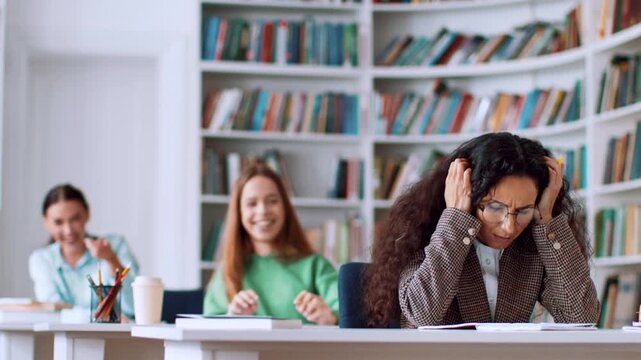 Three students enjoy a moment in a library. Two students smile and laugh while one student focuses on her work. The scene shows a study session filled with friendship.