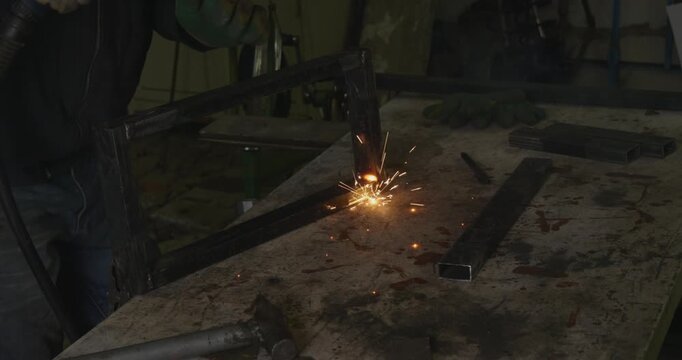 Industrial worker welding a metal frame in a workshop, with bright sparks flying from the electric arc as they join steel profile pipes together on a workbench for fabrication.