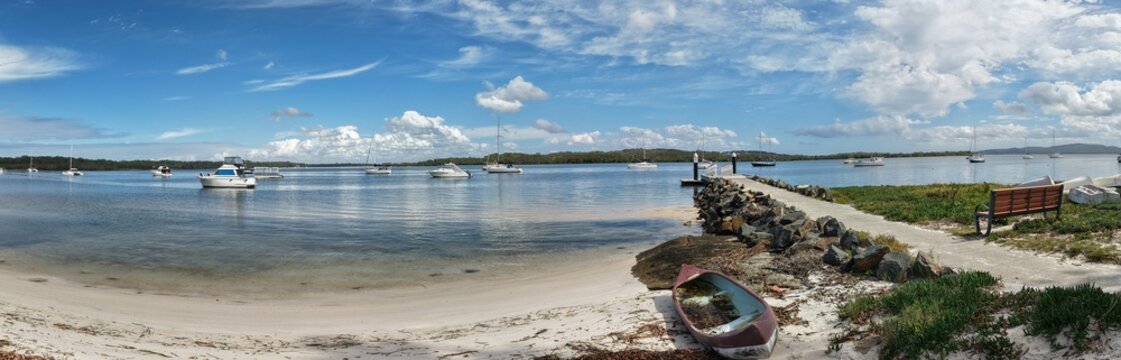 Aerial view of boats bobbing gently on the water, a pier stretches into the calm bay, and a rustic boat sits on the sandy shore, Forster, Australia.