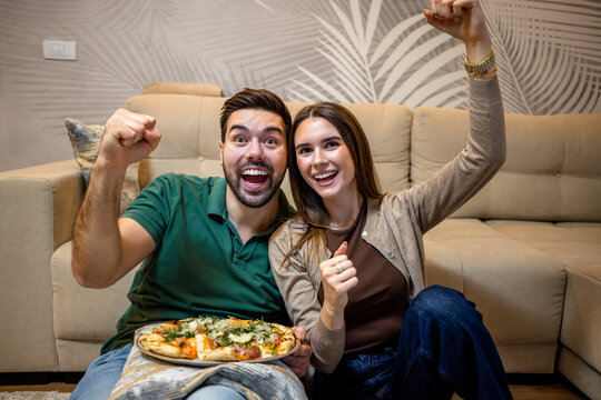 Young couple enjoying pizza and watching a sports match in living room.