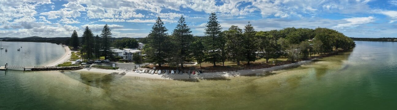 Aerial view of a tranquil peninsula with tall pines casting shadows on the sandy beach, contrasting with the clear turquoise waters, Forster, New South Wales, Australia.
