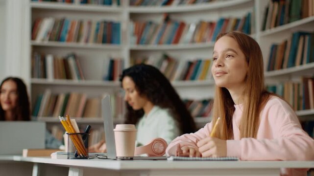 A girl with light skin writes in a notebook while focused on her work. Two other students are nearby working on laptops in a library filled with books.