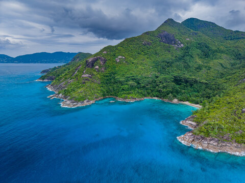 Aerial view of the island's lush, verdant mountains meeting the tranquil, turquoise sea under a clouded sky, Vista do Mar, Mahe Island, Seychelles.