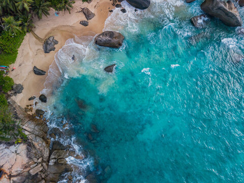 Aerial view of turquoise waves crashing against the sandy shore, fringed by verdant trees and scattered rocks, Vista do Mar, Seychelles.