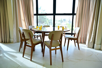 Fototapeta premium round wooden dining table with beige chairs in bright room, elegant interior design for dining area with large window, modern dining room furniture set with yellow table setting, sunlit breakfast nook