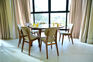 Fototapeta premium round wooden dining table with beige chairs in bright room, elegant interior design for dining area with large window, modern dining room furniture set with yellow table setting, sunlit breakfast nook