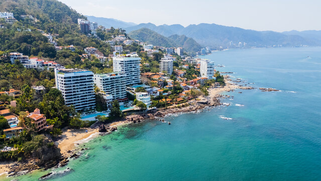 Aerial view of where emerald waters meet the sandy shore, fringed by vibrant green hills dotted with buildings, Puerto Vallarta, Jalisco, Mexico.