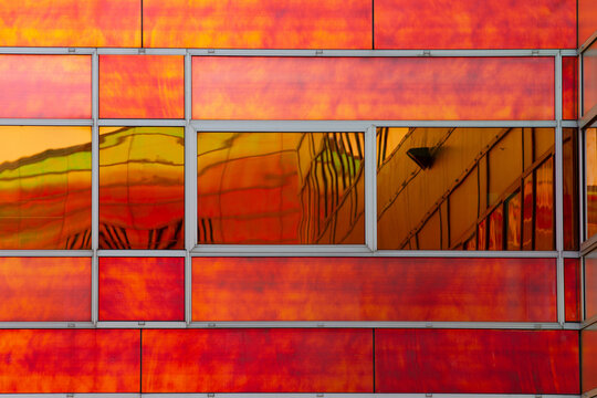 View of a vibrant orange and red facade reflecting a distorted cityscape, creating a surreal and abstract composition, La Defense, Flevoland, Netherlands.