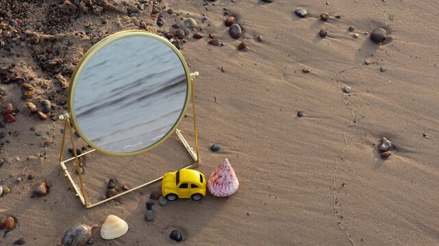 Round mirror with golden frame on sandy beach reflecting sea waves near yellow toy car and seashell on sunny day. Concept of reflection, summer vacation, travel aesthetics and beach lifestyle. 