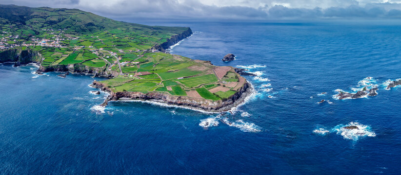 Aerial view of vibrant green fields meet the rugged coastline where the Estrada do Farol winds along the cliffs, Flores, Acores, Portugal.
