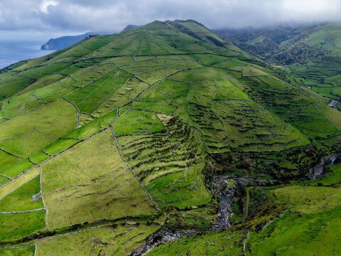Aerial view of emerald pastures quilted across undulating hills, bisected by a winding stream, creating a vibrant tapestry of nature, Flores, Acores, Portugal.