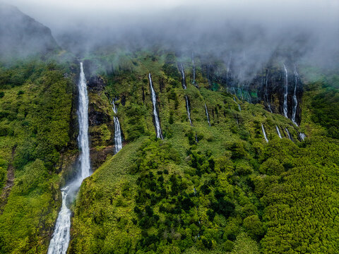 Aerial view of multiple waterfalls cascading down a verdant cliff face, shrouded in mist, creating a serene, ethereal landscape, Flores, Azores, Portugal.