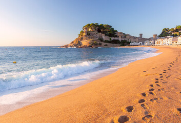 Obraz premium Golden morning view of the sandy beach and fortified old town of Tossa de Mar, Spain, with gentle Mediterranean waves and warm sunrise light along the Costa Brava coast.