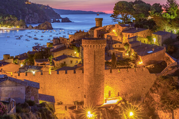 Elevated night view of the fortified old town of Tossa de Mar, Spain, with illuminated stone walls, towers, and the calm Mediterranean bay under deep blue twilight.