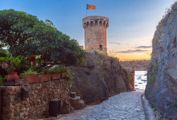 Historic stone watchtower overlooking the calm bay of Tossa de Mar, Costa Brava, Spain, with anchored boats and warm sunset light over the Mediterranean coast.