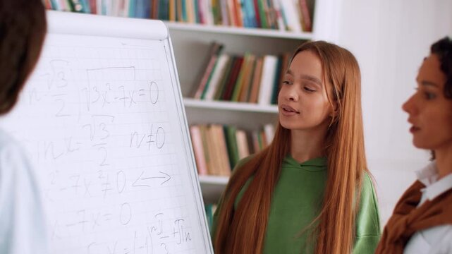 A woman is presenting math equations on a whiteboard while a girl listens attentively. They are inside a classroom with bookshelves filled with books.