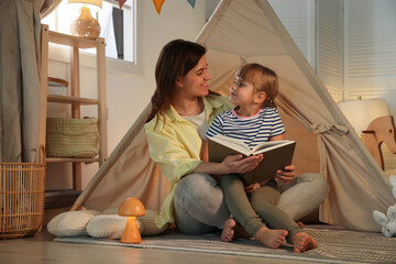Mother and her daughter reading book near toy wigwam at home © New Africa