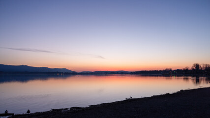 Fototapeta premium Calm lake at sunset with soft pastel sky, distant hills and shoreline silhouettes reflected in still water.
