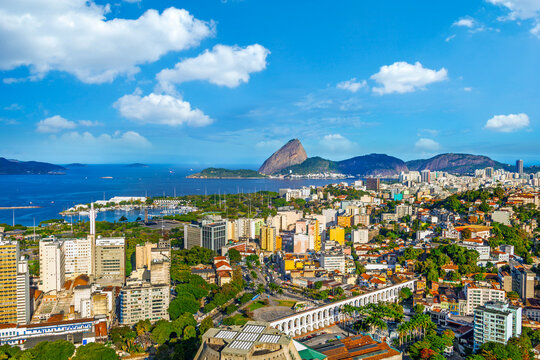 Panoramic aerial view of Rio de Janeiro featuring the Lapa Arches, Metropolitan Cathedral, and lush urban neighborhoods leading to Guanabara Bay and the iconic Sugarloaf Mountain under a blue sky.