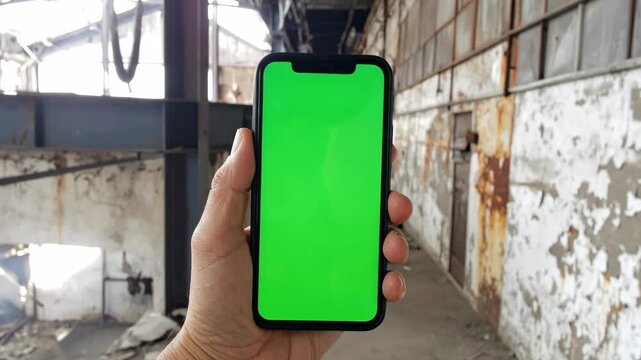 Hand holds iPhone with green screen in abandoned industrial building, ideal for digital content creation and virtual applications.