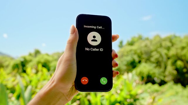 Hand Holding Smartphone with No Caller ID Incoming Call Displayed Against Sunny Grass and Blue Sky, Concept Privacy Risk and Unknown Contact