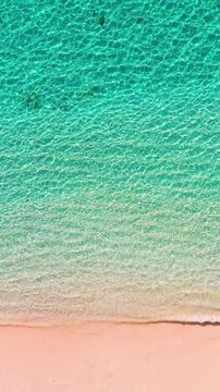 Vertical drone shot of a peaceful tropical beach with calm turquoise water and gentle waves washing onto the sand. Serene coastal landscape, perfect for travel and relaxation content.
