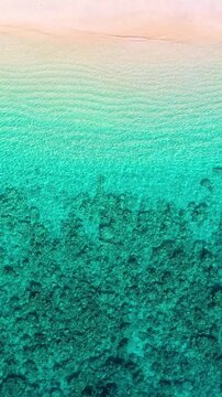 Vertical drone shot of a peaceful tropical beach with calm turquoise water and gentle waves washing onto the sand. Serene coastal landscape, perfect for travel and relaxation content.