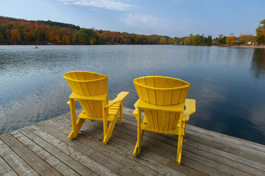 Two bright yellow Muskoka chairs face a wide Ontario lake bay from a weathered wooden dock, with rolling hills of vibrant peak autumn foliage curving around both shores on a partly cloudy fall day.