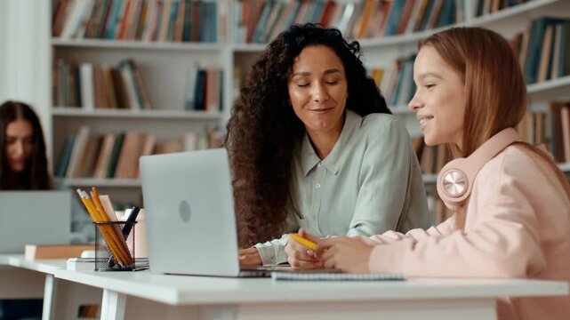In a library, a woman with curly hair sits beside a girl wearing headphones. They focus on a laptop, sharing ideas and working together. The library background features bookshelves filled with books.