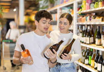 Fototapeta premium Joyful couple of lovers choose dry or sweet wine in a hypermarket, standing in the department with alcohol against the background of customers