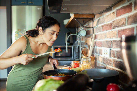Woman tasting vegetable stir fry while cooking in home kitchen